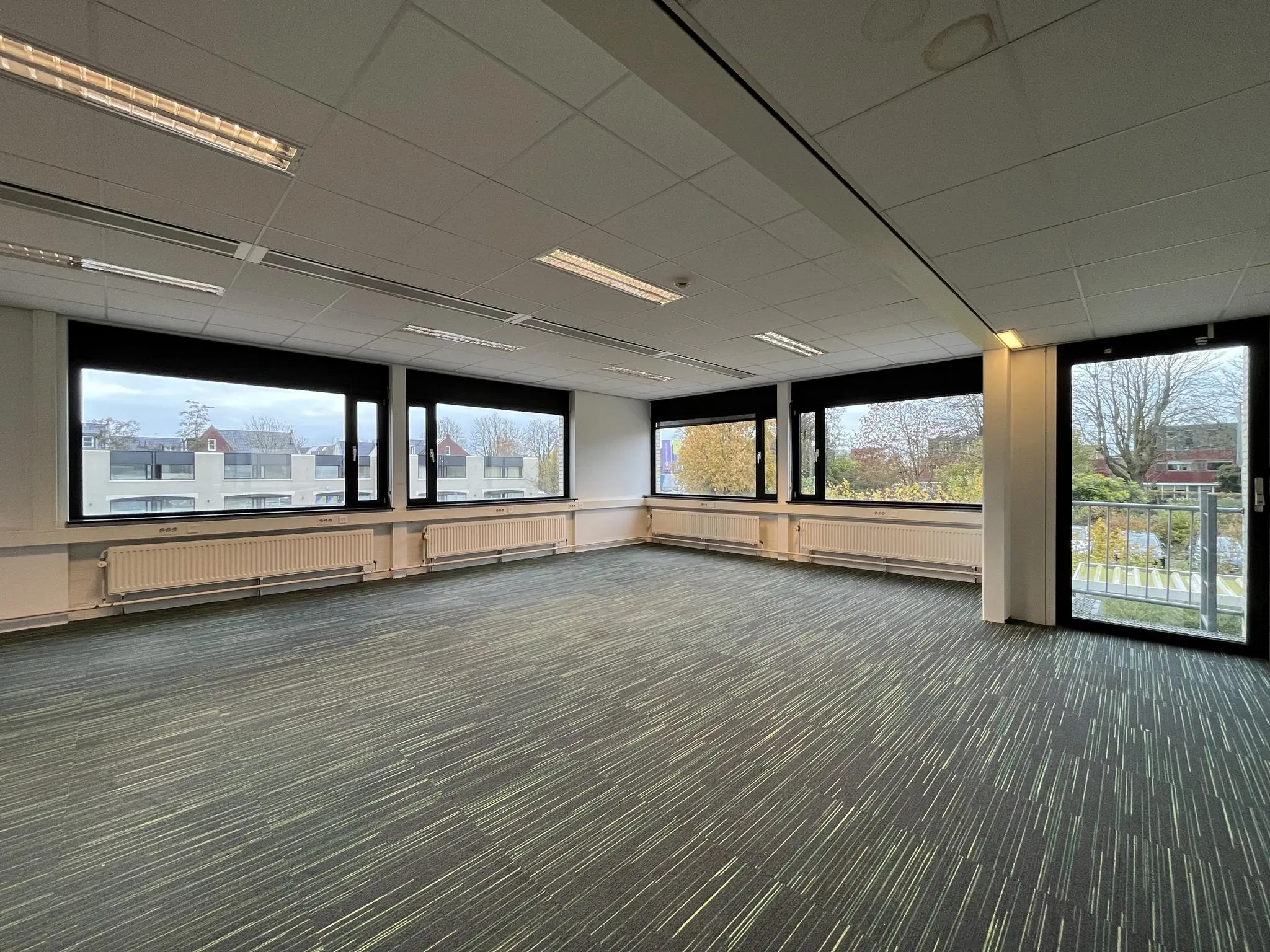 Lichte, lege kantoorruimte aan de Burgemeester Verderlaan met grote ramen en uitzicht op omliggende gebouwen en bomen.